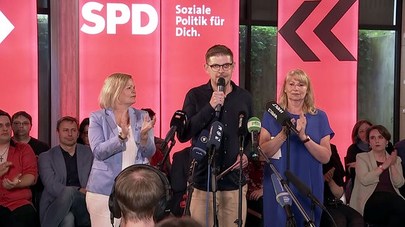 Drei Politiker bei einer Veranstaltung sprechen in Mikrofone.