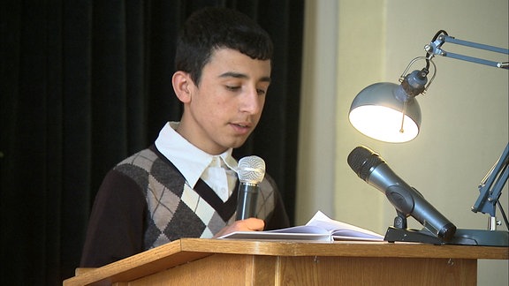 Ein junger Mann steht an einem Rednerpult und liest etwas vor.