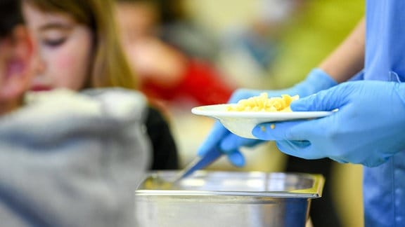 Hände mit blauen Handschuhen geben Essen aus, im Hintergrund Schüler
