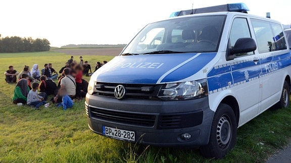 Eine Gruppe von Migranten aus Syrien und der Türkei sitzt neben einem Einsatzfahrzeug der Polizei.