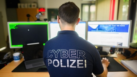 Ein Polizist steht in einem Poloshirt mit Aufschrift "Cyber Polizei" an einem Computer.