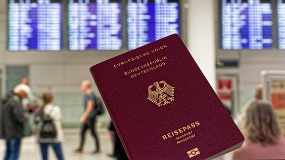 Symbolbild: Eine Hand hält einen deutschen Reisepass vor Flughafenanzeigen.