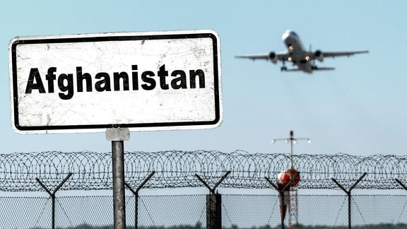 Ein Flugzeug startet hinter einem Stacheldrahtzaun davor ein Schild mit Aufschrift: Afghanistan.