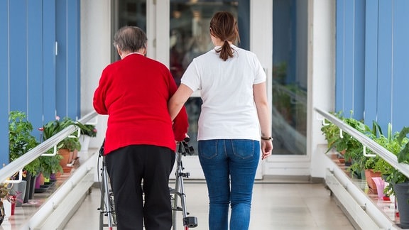 Eine Pflegerin (r) führt eine ältere Frau mit Rollator durch einen Flur in einem Seniorenzentrum.