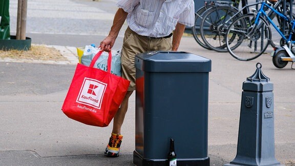 Ein Mann mit Einkaufstaschen in den Händne schaut in einen Mülleimer.