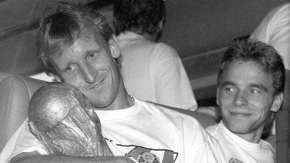 Der deutsche Abwehrspieler Andreas Brehme (l) hält 1990 in Rom den eroberten WM-Pokal im Arm und sein Teamgefährte Thomas Häßler schaut lächelnd zu 