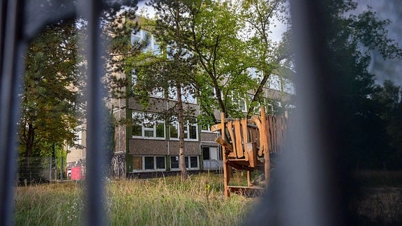 Blick durch einen Zaun auf ein verlassenes Grundstück mit einem Plattenbau. 