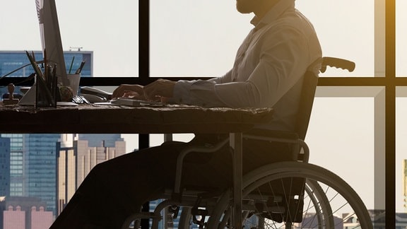 Eine Person sitzt im Rollstuhl an einem Schreibtisch mit Computer.
