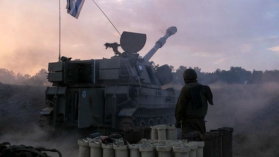 Ein Soldate im Kampfmontur steht vor einem Panzer, auf dem eine israelische Fahne weht.