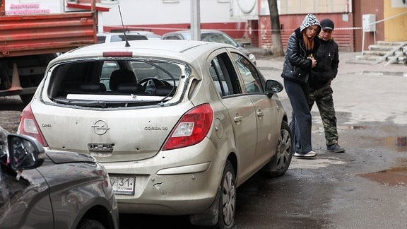 Zwei Menschen gehen an einem, vor einem Wohnhaus geparkten Auto, welches durch ukrainische Militärangriffe beschädigt wurde, vorbei.