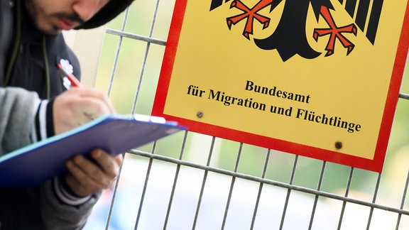 Ein Mann füllt ein Formular aus neben einem Schild vom Bundesamt für Migration und Flüchtlinge an der Landesaufnahmebehörde Niedersachsen am Standort Braunschweig.
