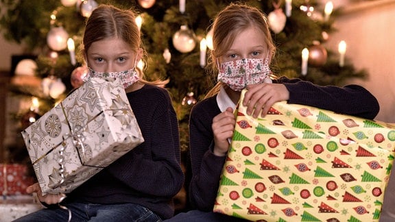 Zwei Kinder mit Masken und Gecshenken vor Weihnachtsbaum