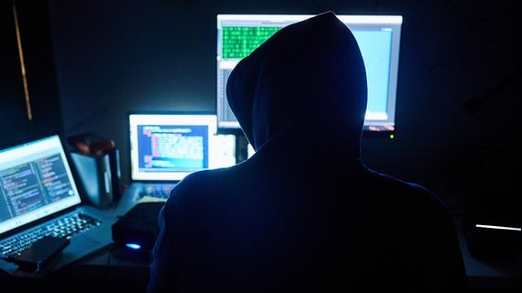 Symbolbild: Fiktive html-Seiten und Hacker-Programme sind auf Bildschirmen zu sehen, während ein Mann seinen Hände auf der Tastatur hat.