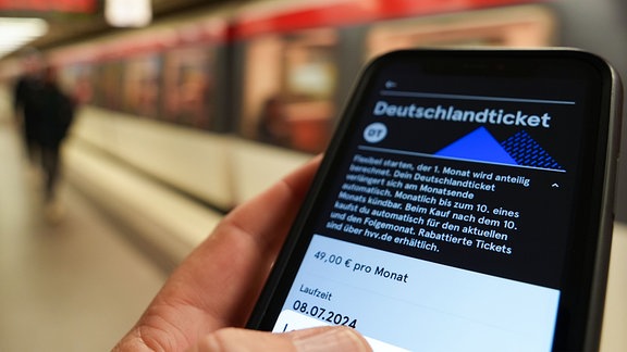 Ein Fahrgast hält ein Smartphone mit einem digitalen Deutschlandticket an einer U-Bahnstation in der Hand.