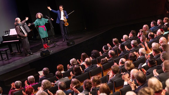 Blick übers applaudierende Publikum in einem Theater hinweg auf die Bühne, wo drei Personen stehen: links ein Mann mit Akkordeon, in der Mitte eine Frau, rechts ein Mann mit Geige.