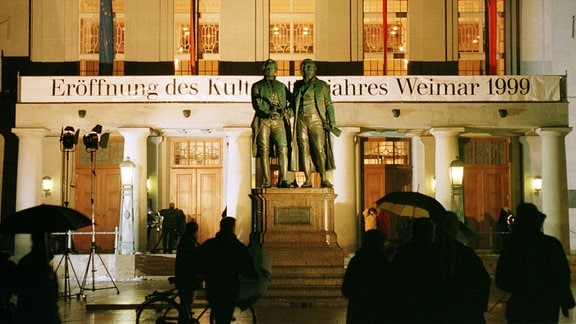 Passanten stehen am Freitag abend (19.02.1999) vor dem hell erleuchteten Deutschen Nationaltheater in der europäischen Kulturhauptstadt Weimar. Im Vordergrund ist das Goethe-Schiller-Denkmal zu sehen. Hier wurde heute das Kulturstadtjahr 1999 offiziell eröffnet. 