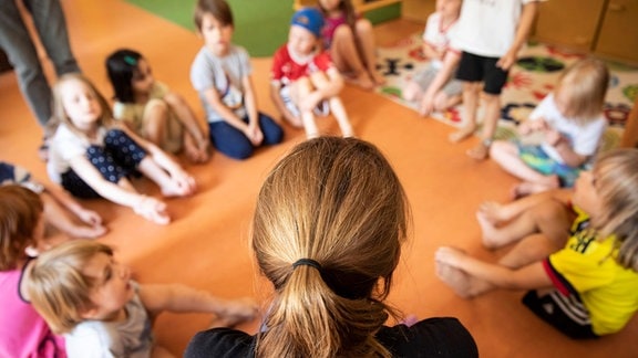 Eine Erzieherin sitzt mit Kindern im Kindergarten im Kreis auf dem Boden