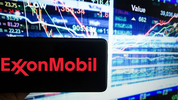 Das Logo von Exxon Mobil ist auf einem Bildschirm von einem Handy neben einer Illustration des schnelllebigen Aktienmarkts zu sehen.