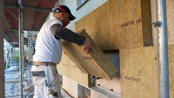 Ein Mitarbeiter einer Baufirma setzt zur Dämmung der Fassade eine Steinwollplatte an einer Hauswand an.