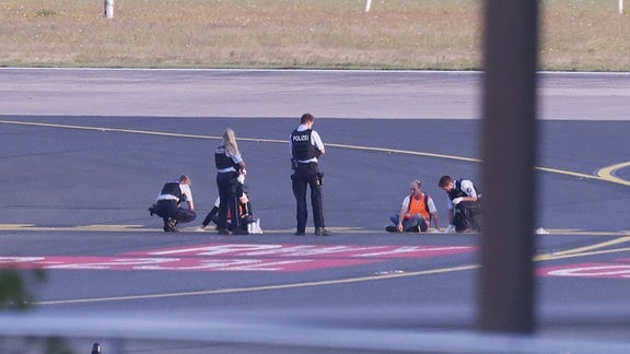 Polizisten stehen auf dem Flugfeld und versuchen Aktivisten der Gruppe Letzte Generation am Flughafen vom Asphalt zu lösen, nachdem sie sich festgeklebt haben.