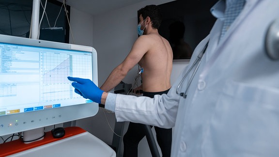 Arzt analysiert Daten auf einem Monitor während ein Patient auf einem Laufband geht.