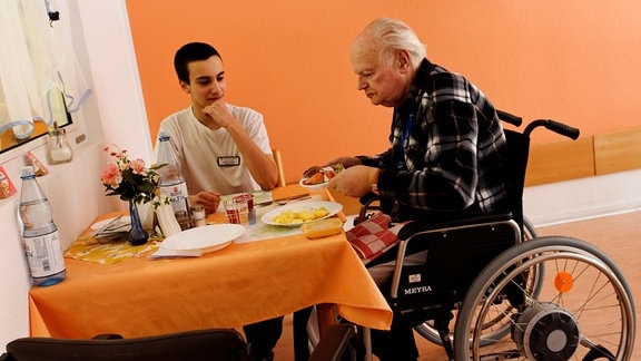 Ein Bundesfreiwilligendienst-Leistender betreut einen Senior im Rollstuhl beimEssen.