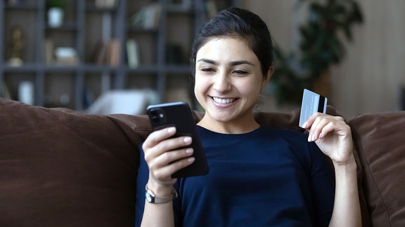 Lächelnde junge indische Kundin beim Online-Einkauf mit Smartphone und Debitkarte.  