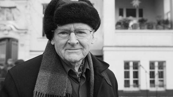 Schriftsteller Gerhard Wolf, mit einer Fellmütze, Mantel und Schal tragend vor einem Altbau