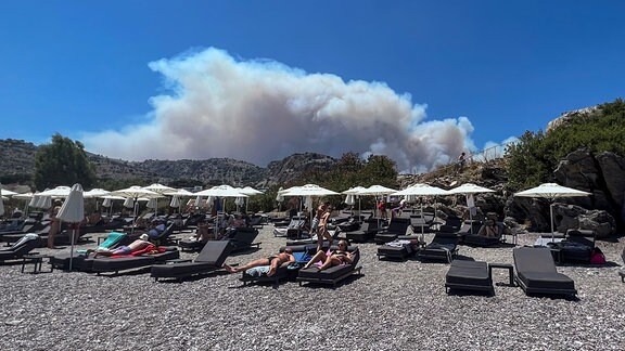 Am Viglika Beach auf der griechischen Insel Lindos aus, wo sich Menschen auf Sonnenliegen am Strand aufhalten, kann man aufsteigenden Rauch von Waldbränden auf der Ferieninsel Rhodos sehen.