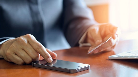 Ein schwarzes Smartphone und eine Kreditkarte in den Händen eines Mannes vor weißen Hintergrund.