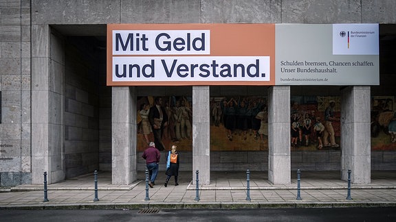 Über dem Eingang zum Bundesministerium der Finanzen in Berlin hängt ein Plakat mit der Aufschrift „Mit Geld und Verstand. Schulden bremsen, Chancen schaffen. Unser Bundeshaushalt.“; darunter laufen zwei Personen in Richtung des Eingangs des Ministeriums