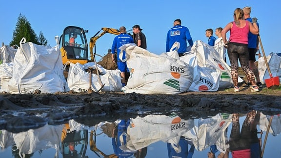 Eine Gruppe Menschen, die mit Hilfe eines kleinen Baggers Sandsäcke befüllt, spiegelt sich im Wasser.