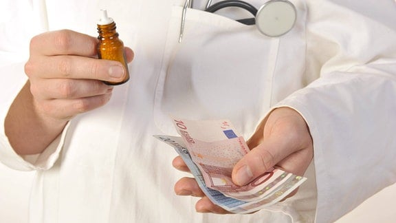 Ein Arzt hält Geldscheine und homöopathisches Medikament in der Hand.