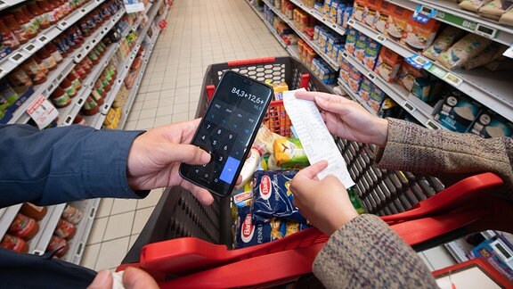 Vor einem vollen Einkaufswagen rechnen Zwei Personen mit einem Taschenrechner und halten einen Bon