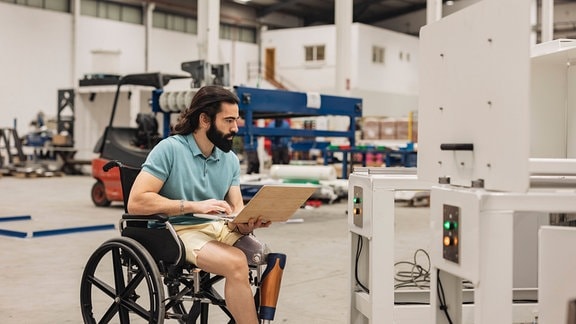 Ein Ingenieur im Rollstuhl sitzend, der in einer Fabrik eine Maschine über einen Laptop analysiert.