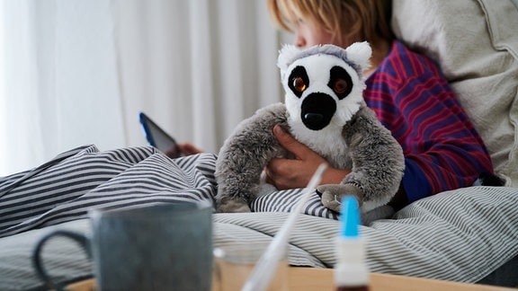 Ein Fieberthermometer, Nasenspray und eine Tasse Tee stehen vor einem Bett, in dem ein Kind mit Kuscheltier sitzt und auf ein Tablet schaut.