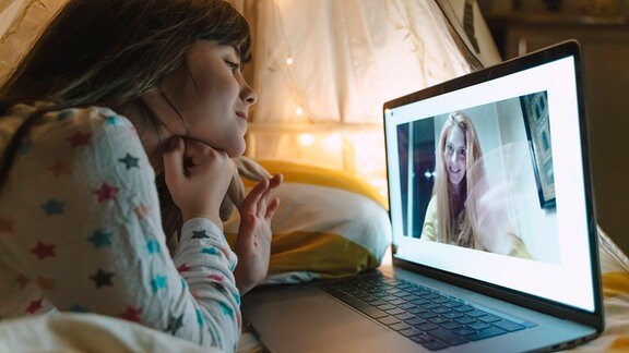 Ein Kind liegt im Bett vor einem Laptop und spricht mit einer Frau im Videochat.