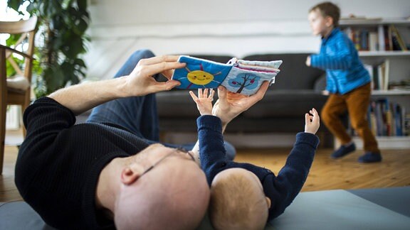 Ein Vater spielt mit seinem Baby waehrend im Hintergrund ein zweites Kind tobt.
