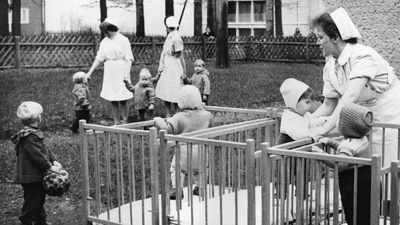 Unter der Obhut von drei Betreuerinnen genießen einige Kleinkinder auf einer Wiese und in fahrbaren Kinderbettchen vor dem Kinder-Betreuungsheim der Heilstätte Bad Berka im November 1960 die frische Luft. 