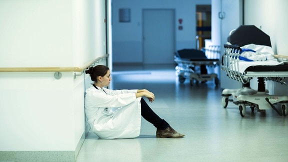 Überforderter junger Arzt sitzt auf dem Krankenhausflur.