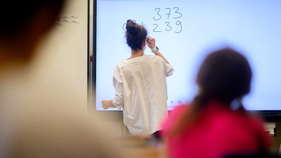 Ein Lehrerin schreibt eine Mathematikaufgabe auf eine digitale Schultafel im Klassenraum einer 4. Klasse einer Grundschule.