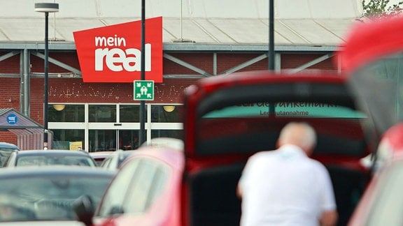 Ein Mann steht zwischen zwei geparkten Autos an einem geöffneten Kofferraum, im Hintergrund ist das Logo der SB-Warenhauskette Mein Real zu sehen.