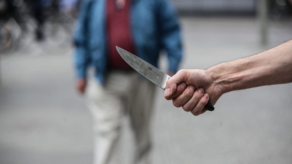 Eine Hand hält drohend ein Messer. Im Hintergrund läuft eine Person. 