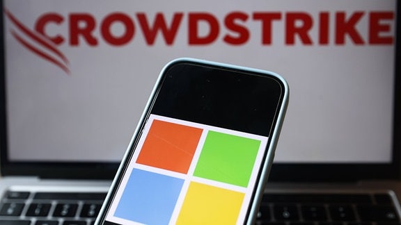Illustration -  Microsoft-Logo auf einem Telefonbildschirm und das Crowdstrike-Logo auf einem Bildschirm im Hintergrund