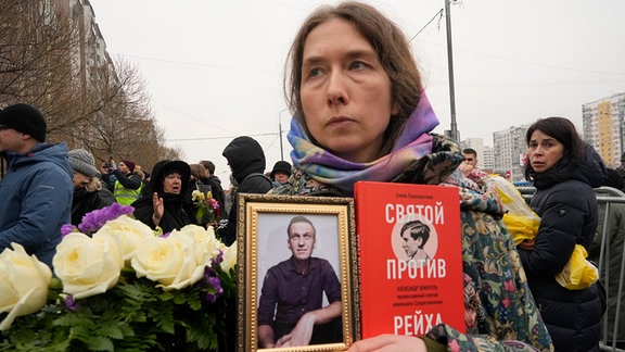 Eine Frau hält ein Porträt von Alexej Nawalny und ein Buch mit dem Titel «Ein Heiliger gegen das Reich» in der Hand, während sich Menschen vor Kirche zu Ehren der Gottesmutterikone «Lindere meine Trauer» im südöstlichen Bezirk Marjino zur Beisetzung des Kremlgegners versammeln.