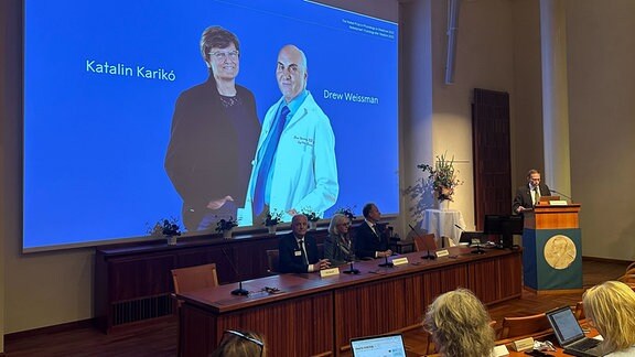 Fotos der Wissenschaftlerin Katalin Kariko und des Wissenschaftlers Drew Weissman sind bei der Bekanntgabe des Nobelpreises für Medizin auf einer Leinwand zu sehen