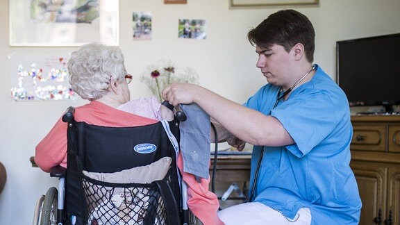 Eine maennliche Pflegekraft misst Blutdruck bei einer aelteren Frau im Rollstuhl.