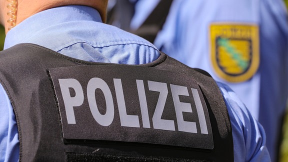 Aufschrift Polizei auf Polizeiweste im Anschnitt. Im Bildhintergrund das Landeswappen Polizei Sachsen in der Bildunschärfe.