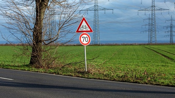 Ein Verkehrsschild mit dem Hinweis auf das Tempolimit 70 und "Achtung, Radfahrer" steht an einer Landstraße. Dahinter stehen zahlreiche Strommasten auf Feldern.