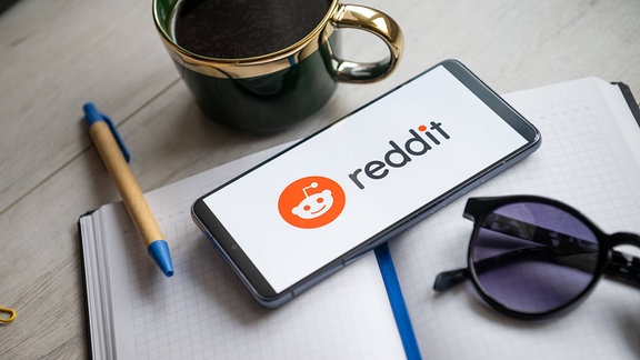 Ein Smartphone auf dessen Bildschirm das Reddit Logo zu sehen ist, liegt auf einem Tisch mit einem Notzibuch und einer Kaffeetasse.